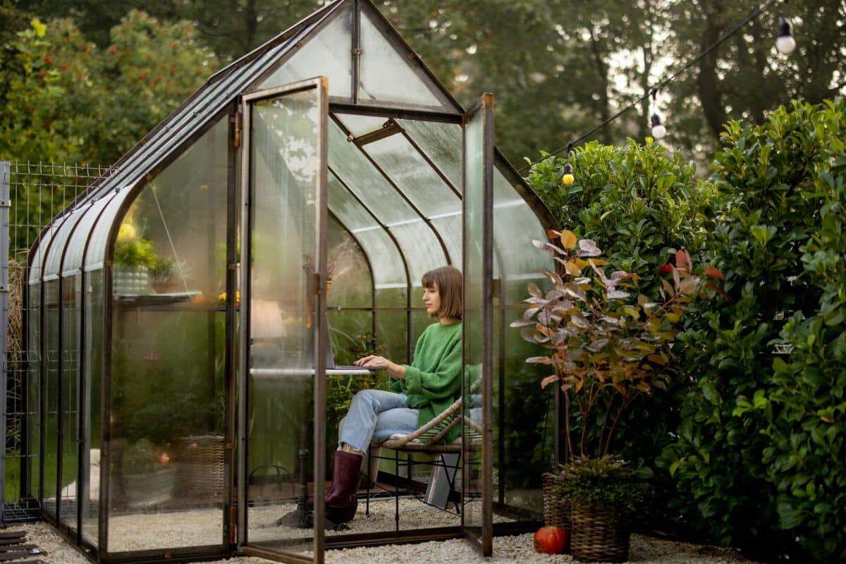 Woman works remotely on laptop in glasshouse at backyard